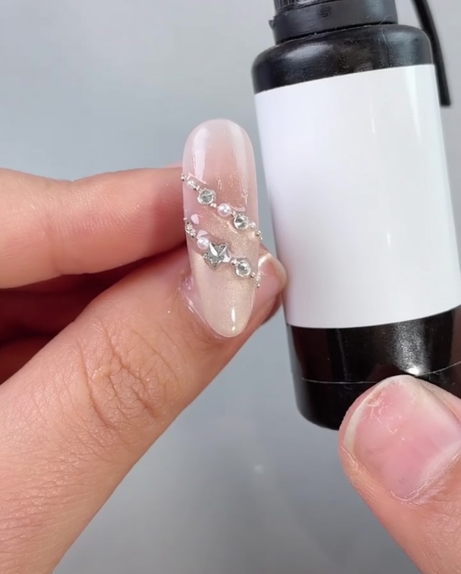 Un pincel de uñas aplicando un top coat o sellador transparente sobre una uña decorada con perlas y diamantes.