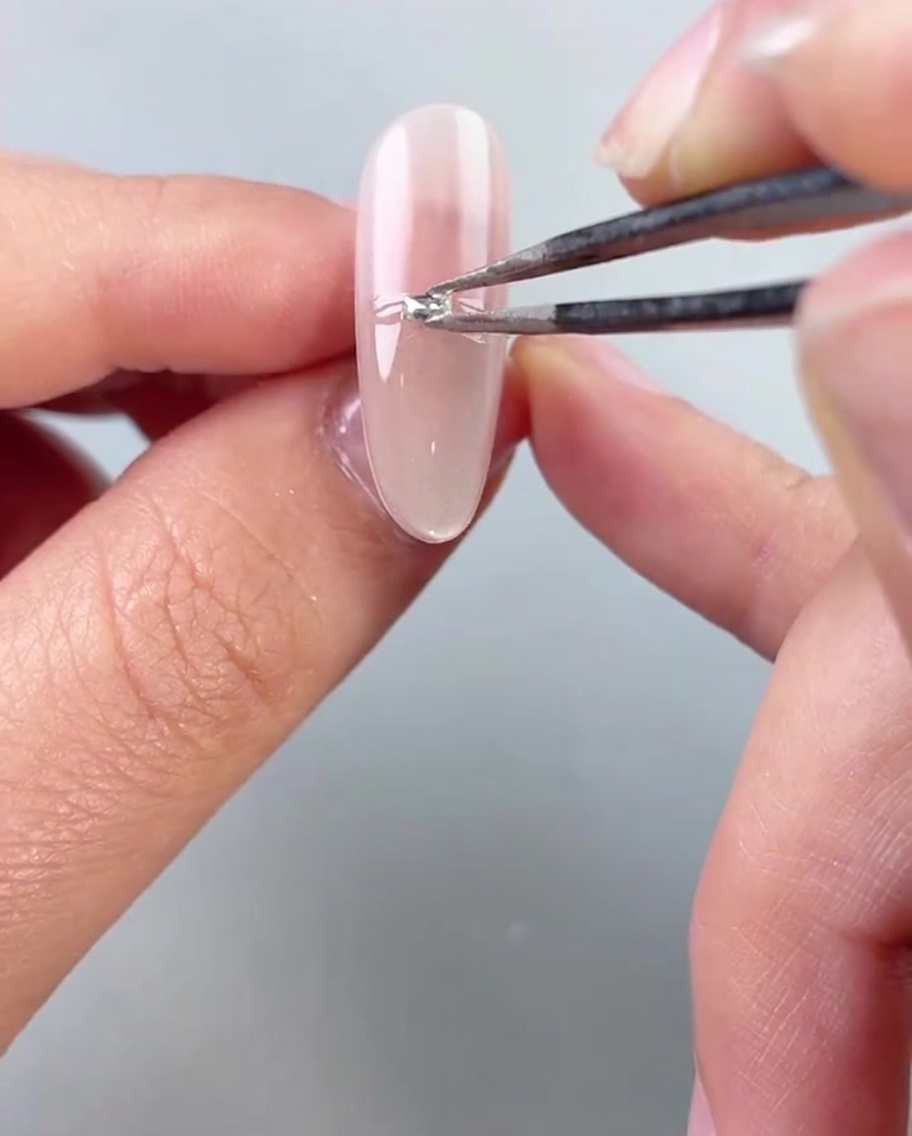 Unas pinzas colocando pequeñas cuentas o diamantes plateados horizontalmente a través de la mitad de una uña pintada con gel.