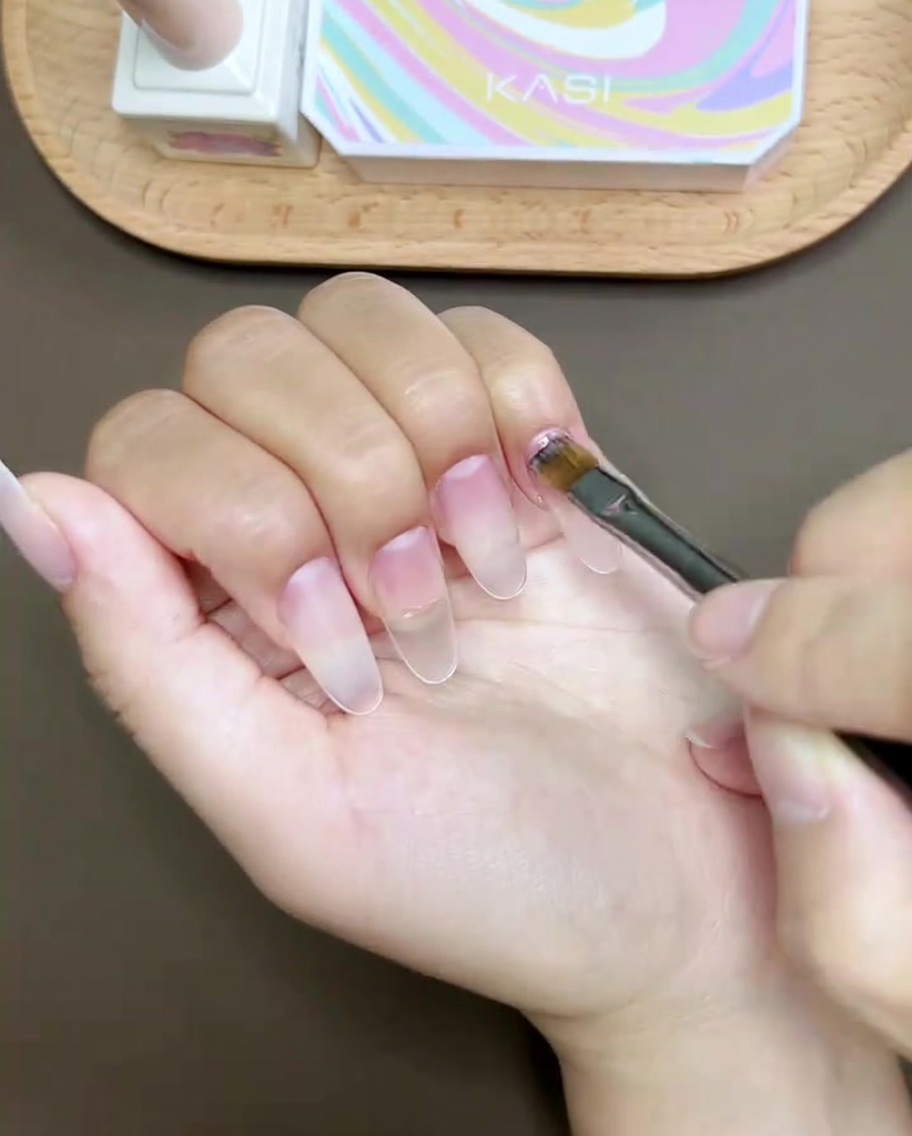 Un pincel de uñas pintando una línea curva rosa a través de la punta de la uña de un pulgar.