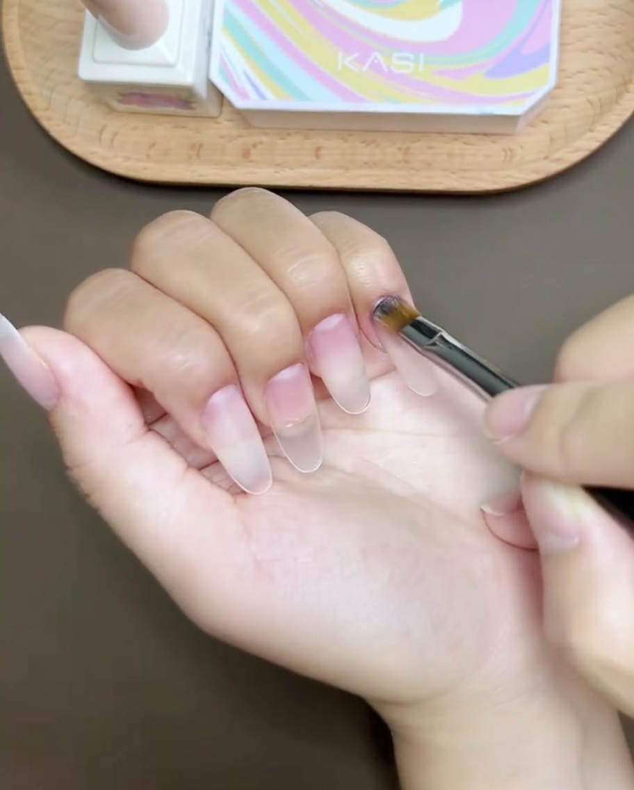 Un pincel aplicando gel de refuerzo transparente sobre una superficie de uña preparada, creando un acabado suave y nivelado.