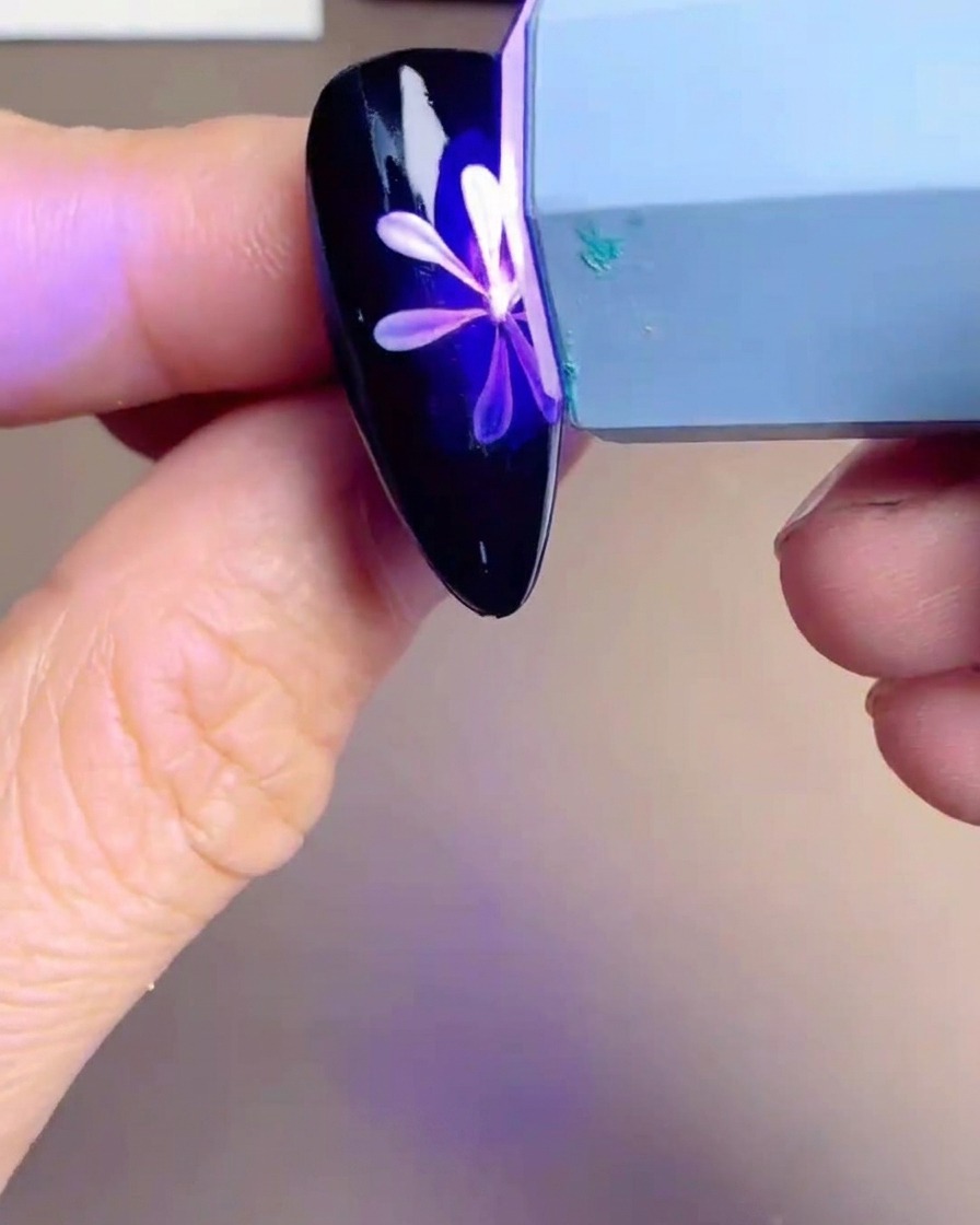 Lámpara UV/LED de mano sobre un tip de uña con un diseño de margarita, mostrando un resplandor púrpura.