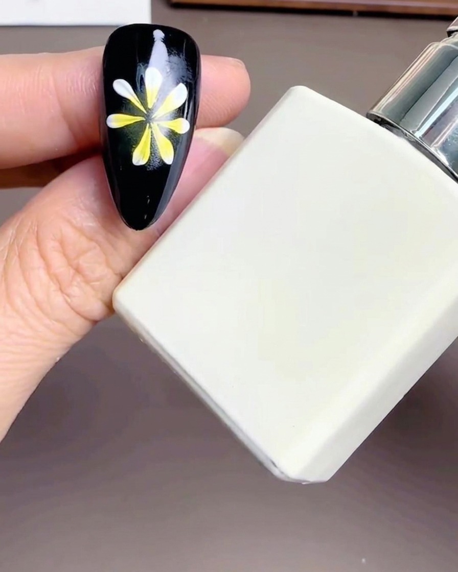 Primer plano de un frasco rectangular blanco de top coat sostenido cerca de una uña con diseño floral.