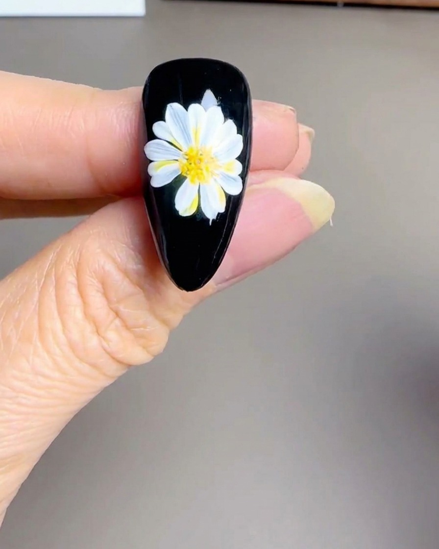 Primer plano de una mano sosteniendo una lámpara UV/LED pequeña sobre una uña negra terminada con diseño de margarita.