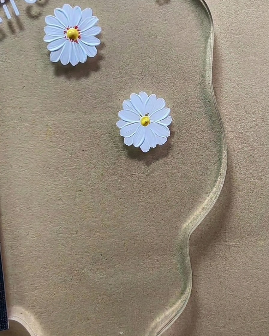 Un pincel de detalle fino aplicando pequeños puntos de color marrón rojizo al centro amarillo de una margarita pintada de blanco sobre una paleta acrílica transparente.