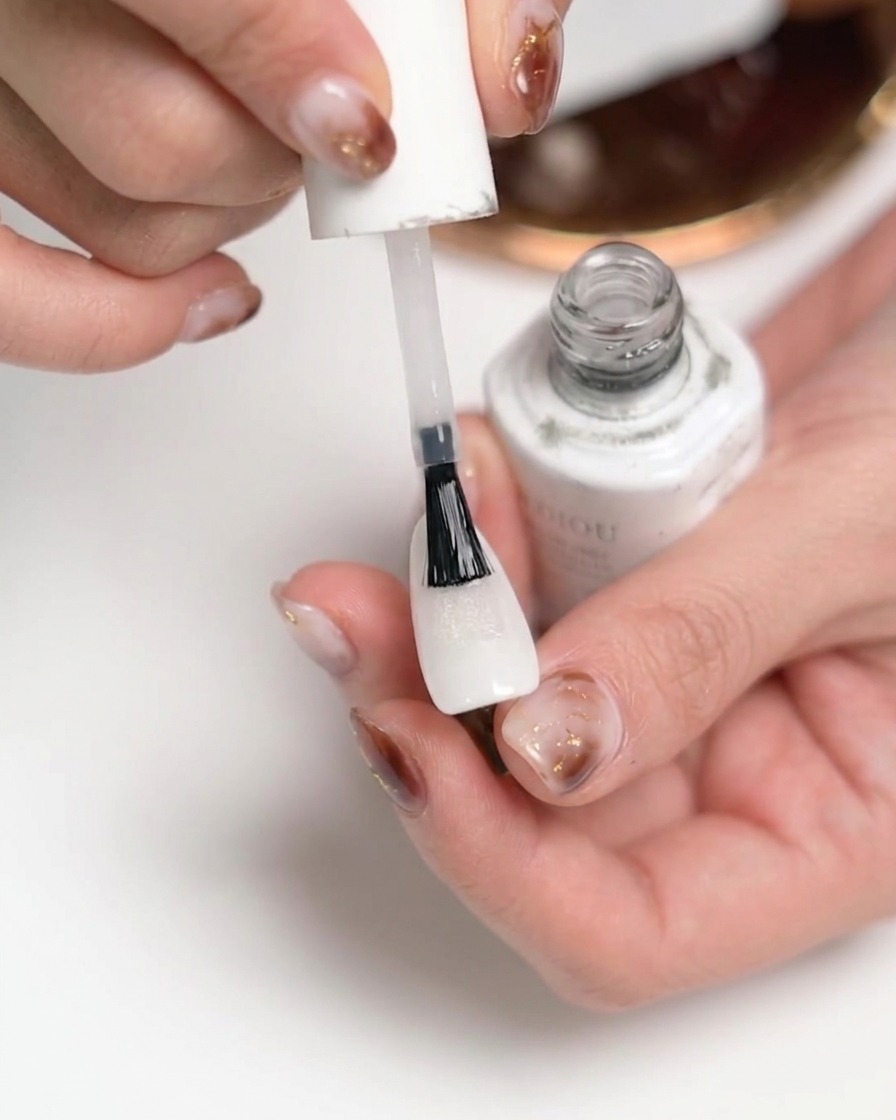 Primer plano de una mano aplicando un top coat mate transparente sobre una uña pintada de blanco con un pincel de un frasco de esmalte blanco.