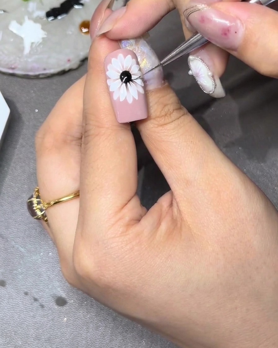 Primer plano de una uña con un diseño floral blanco, donde un pincel fino está haciendo pequeños trazos amarillos hacia afuera desde el centro hacia los espacios entre los pétalos blancos.