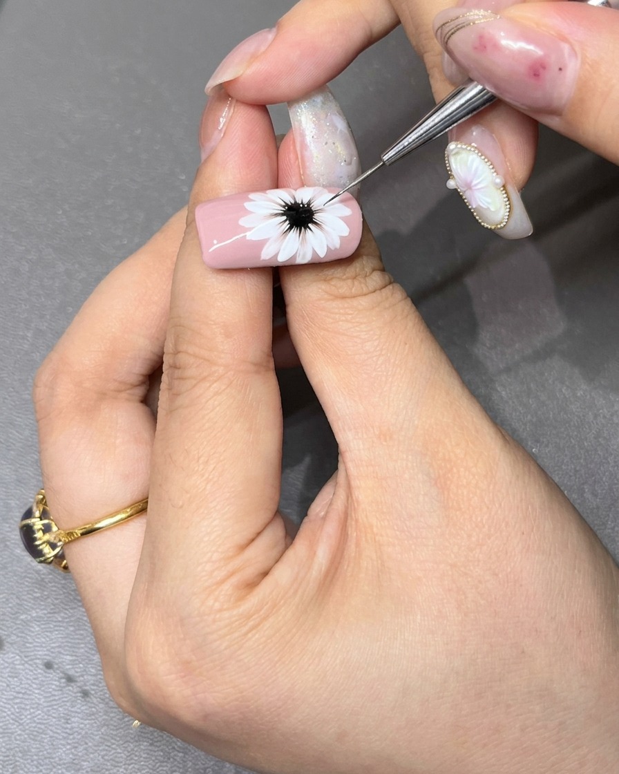 Un pincel de detalle fino refinando el centro negro oscuro de una margarita blanca pintada sobre una uña rosa clara.