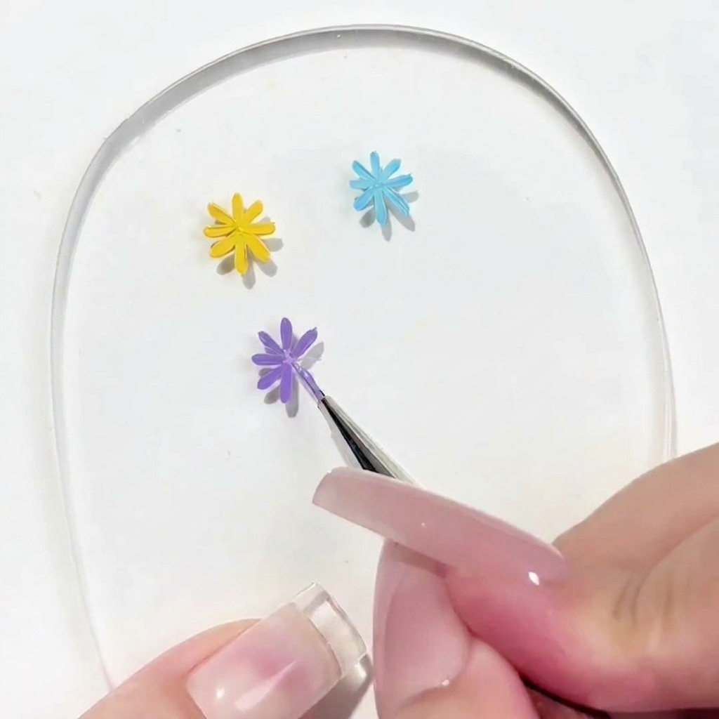 Un primer plano de un pincel de detalle aplicando un pétalo a una margarita morada parcialmente completada en una paleta transparente de nail art.
