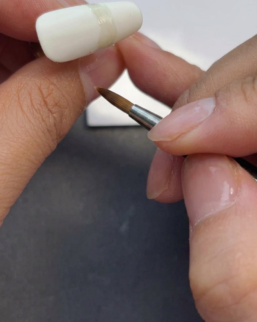 Técnico de uñas aplicando una capa de top coat mate sobre una uña blanca lechosa.