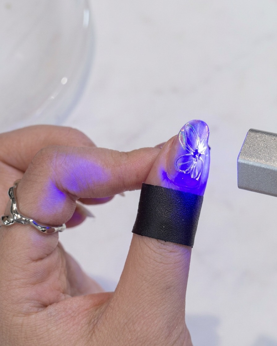 Un pincel aplicando una capa final de top coat transparente sobre una uña con diseño de flor blanca y centro dorado.