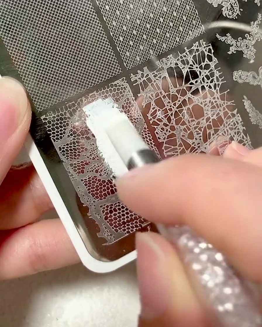 Primer plano de una placa metálica con un diseño de encaje siendo cubierto con gel de estampado blanco.