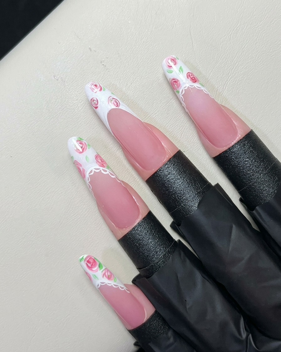 Un pincel de detalle fino pintando hojas verdes diminutas alrededor de rosas rosas sobre una punta francesa de gel blanco.