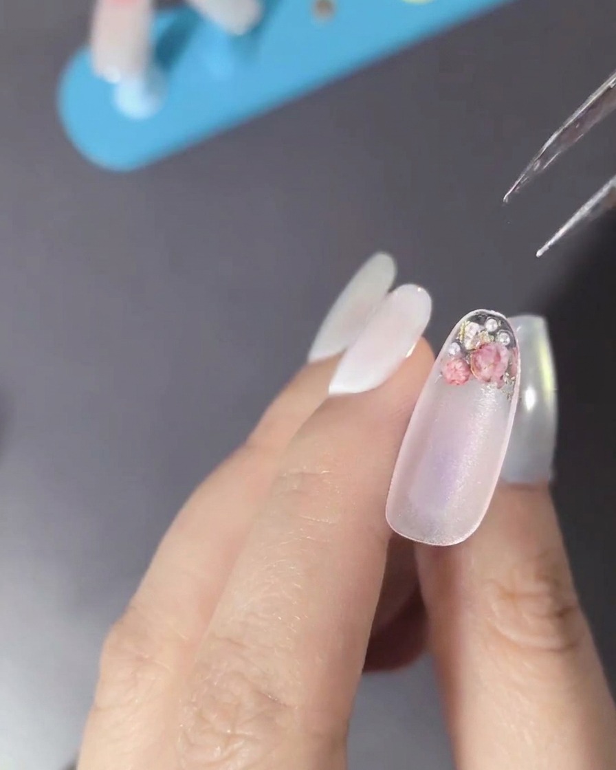 Primer plano de una uña decorada con flores secas, perlas y pequeñas pedrerías metálicas.