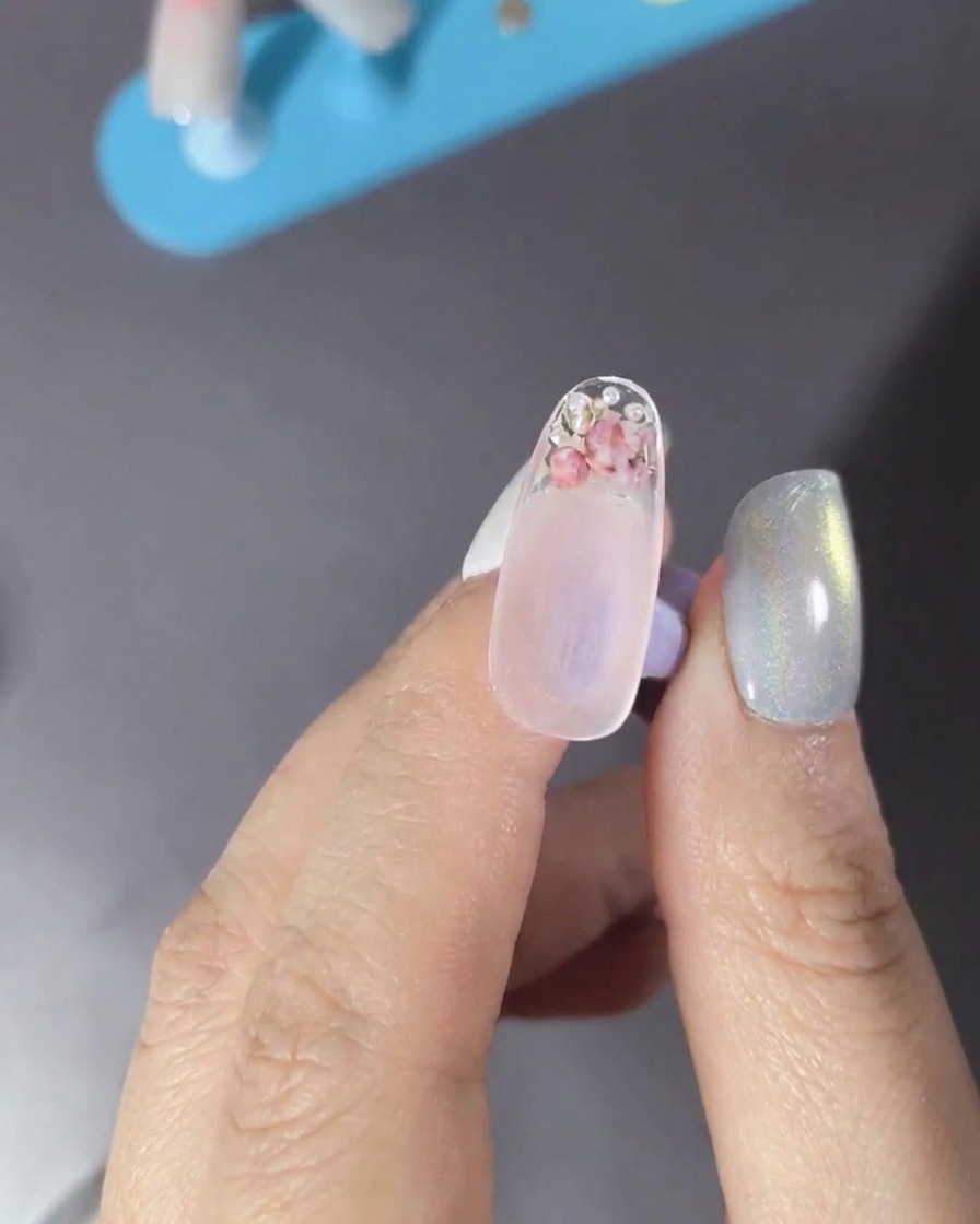 Un pincel aplicando un top coat transparente sobre una uña decorada con flores y perlas.
