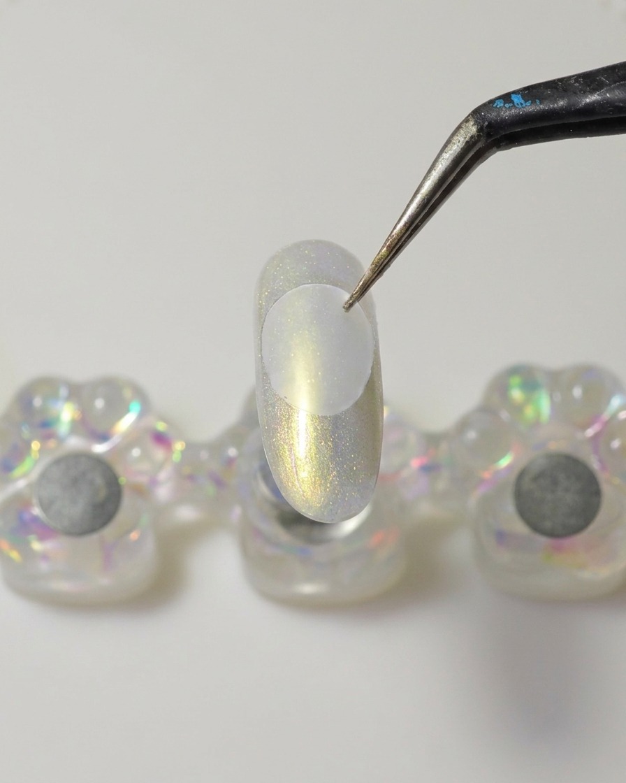 Una espátula metálica aplicando una gota de gel constructor sólido espeso y transparente en la parte inferior de un disco de plástico transparente.