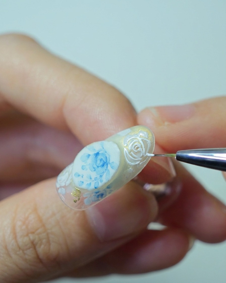 Primer plano de un pincel aplicando gel de relieve blanco espeso sobre los contornos de rosas pintados anteriormente para construir textura 3D.