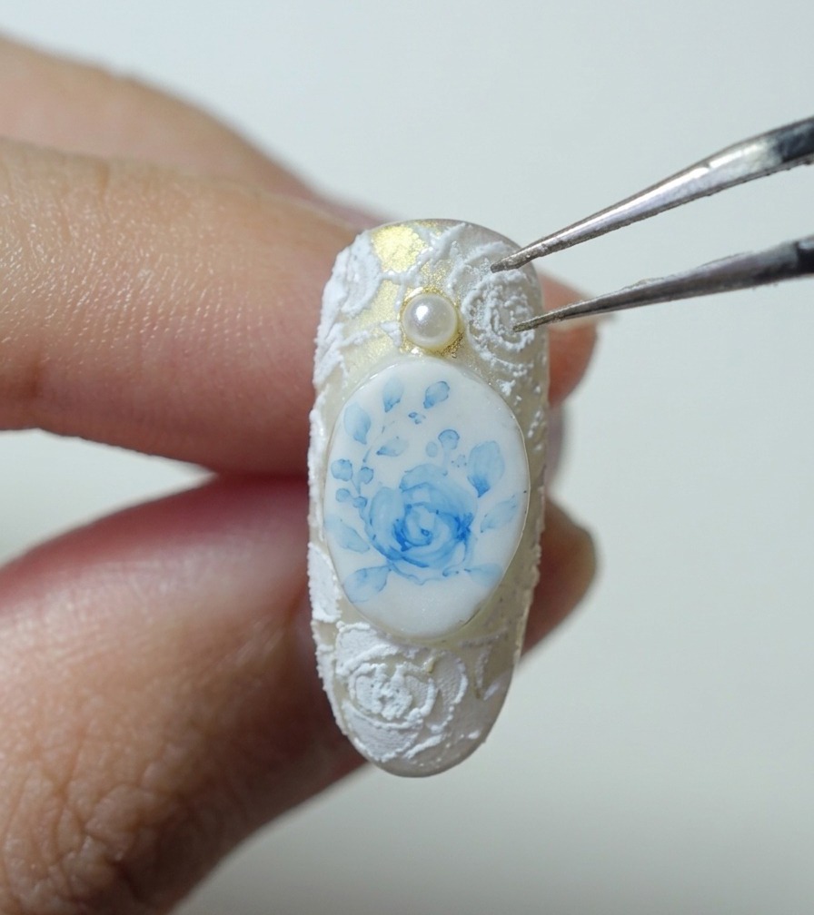 Pinzas sosteniendo una pequeña cuenta de perla blanca sobre una uña con arte de rosa azul y relieve de encaje blanco.