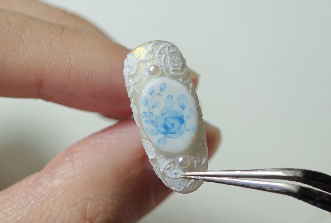 Un primer plano de una uña decorada con rosas azules y encaje blanco, siendo sostenida por pinzas, lista para el curado final.