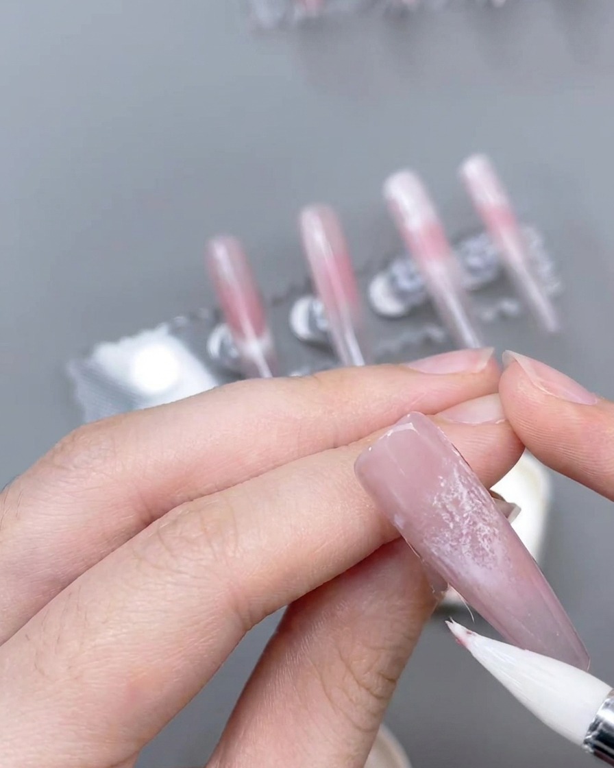 Un pincel delineador fino creando líneas de acento rosa finas y precisas sobre una uña decorada.