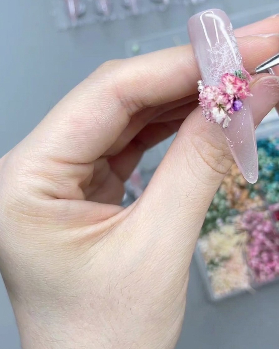 Primer plano de una mano usando una herramienta de precisión para colocar una pequeña perla junto a un grupo de flores secas rosas en una uña postiza larga.