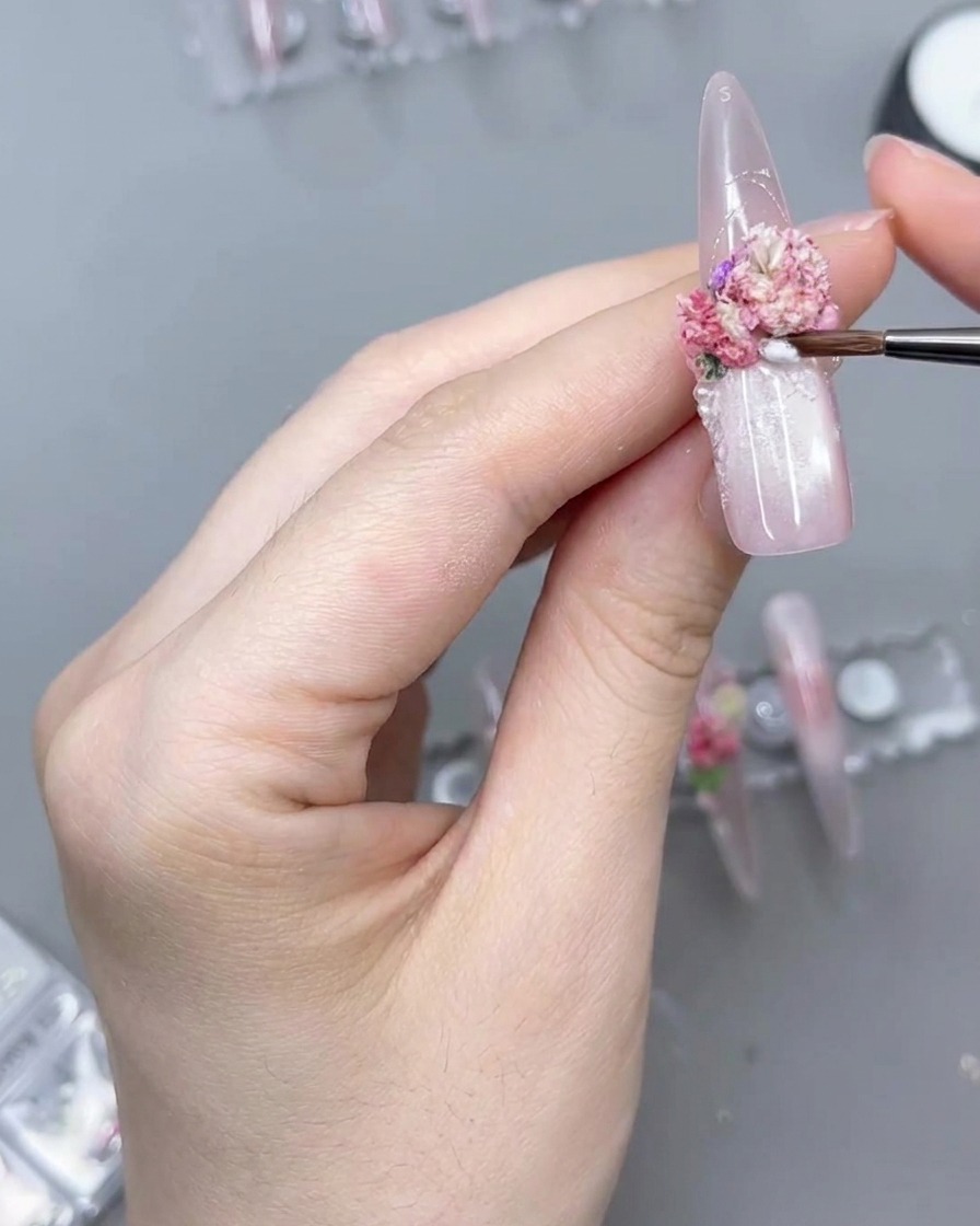 Primer plano de un pincel de detalle fino aplicando producto transparente alrededor de la base de un grupo floral rosa en una uña postiza larga.
