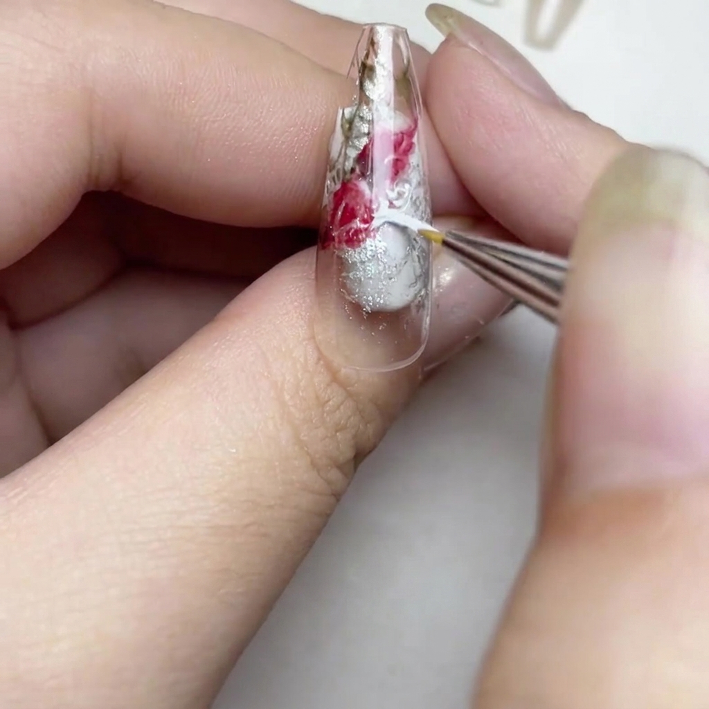 Primer plano de una uña mostrando un pincel añadiendo finos trazos de pintura blanca para crear un efecto de rosa en acuarela suave en los espacios transparentes.