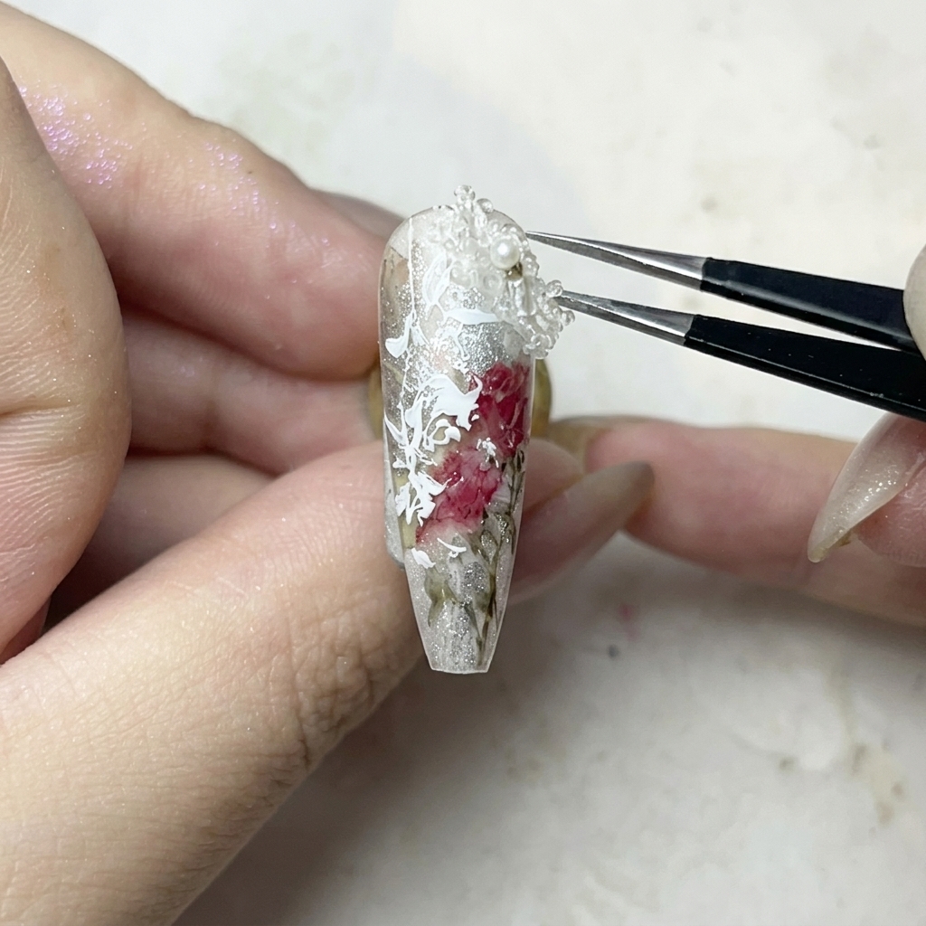 Un par de pinzas colocando cuidadosamente un pequeño dije de flor blanca 3D sobre un punto de pegamento de gel transparente en una uña manicurada.