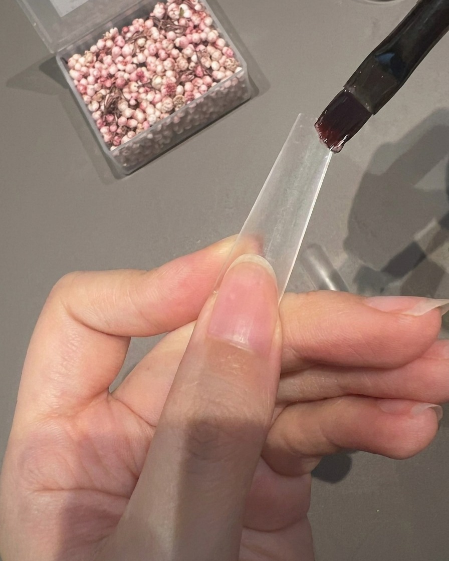 Un pincel aplicando una capa de gel adhesivo transparente en la mitad inferior de una punta de uña transparente.