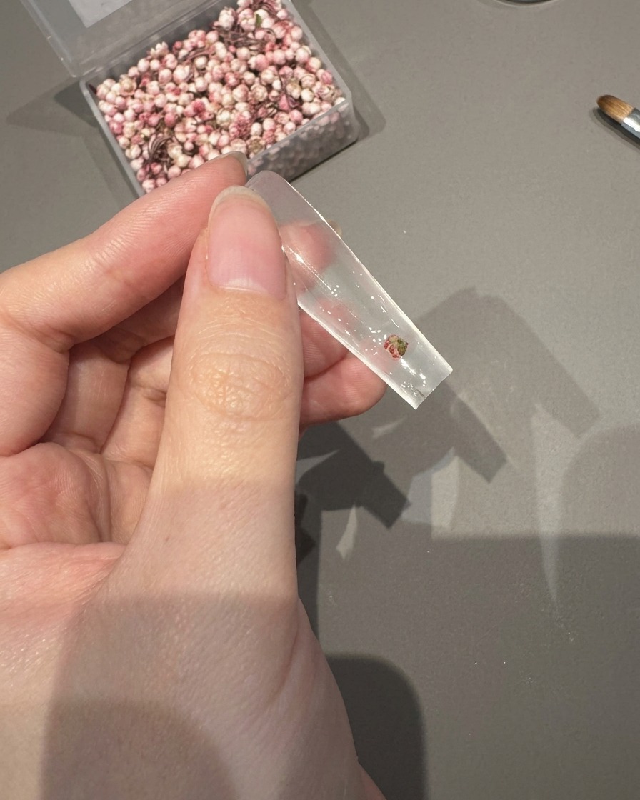 Unas pinzas colocando una pequeña flor seca sobre una punta de uña transparente recubierta de gel adhesivo húmedo.