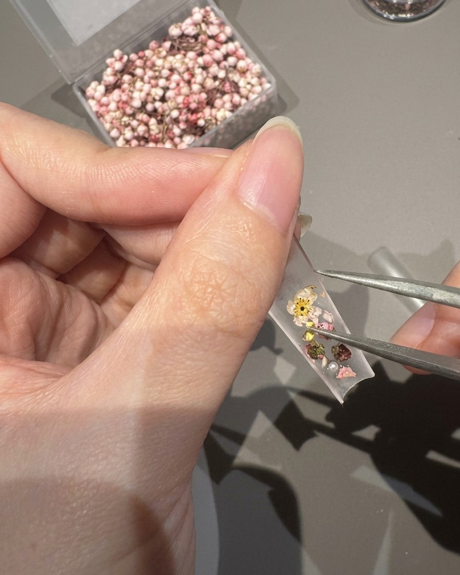 Primer plano de unas pinzas colocando pequeñas perlas y cristales sobre una punta de uña decorada con flores secas.