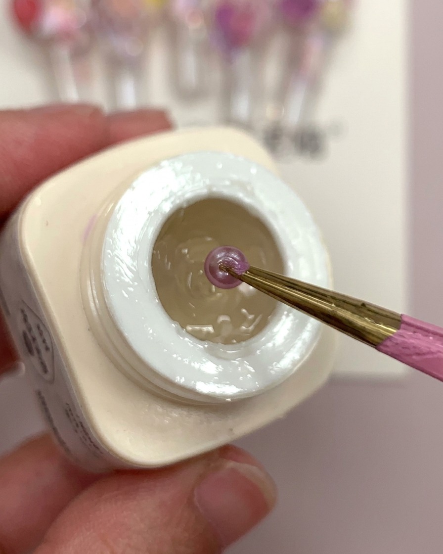 Una mano sosteniendo un tip de uña transparente con una gota de pegamento escarchado y bolas florales moradas siendo colocadas en él.
