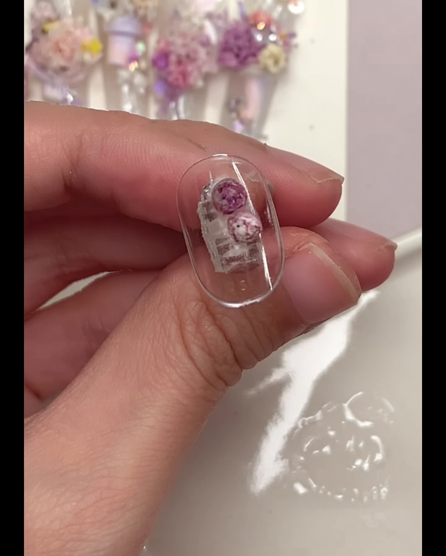 Un pincel de detalle presionando suavemente bolas florales moradas colocadas sobre un tip de uña transparente.