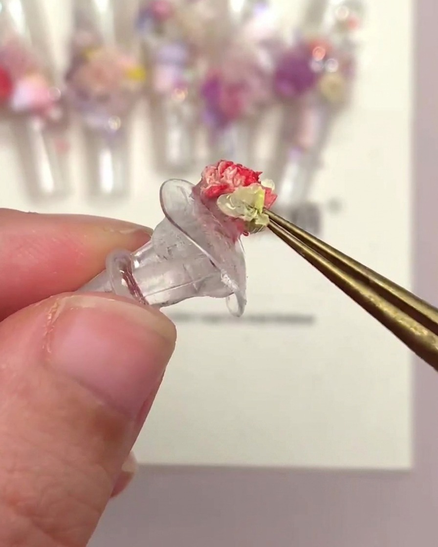 Una mano usando pinzas para colocar una cabeza de flor en forma de trompeta sobre un tip de uña decorado con otras flores.