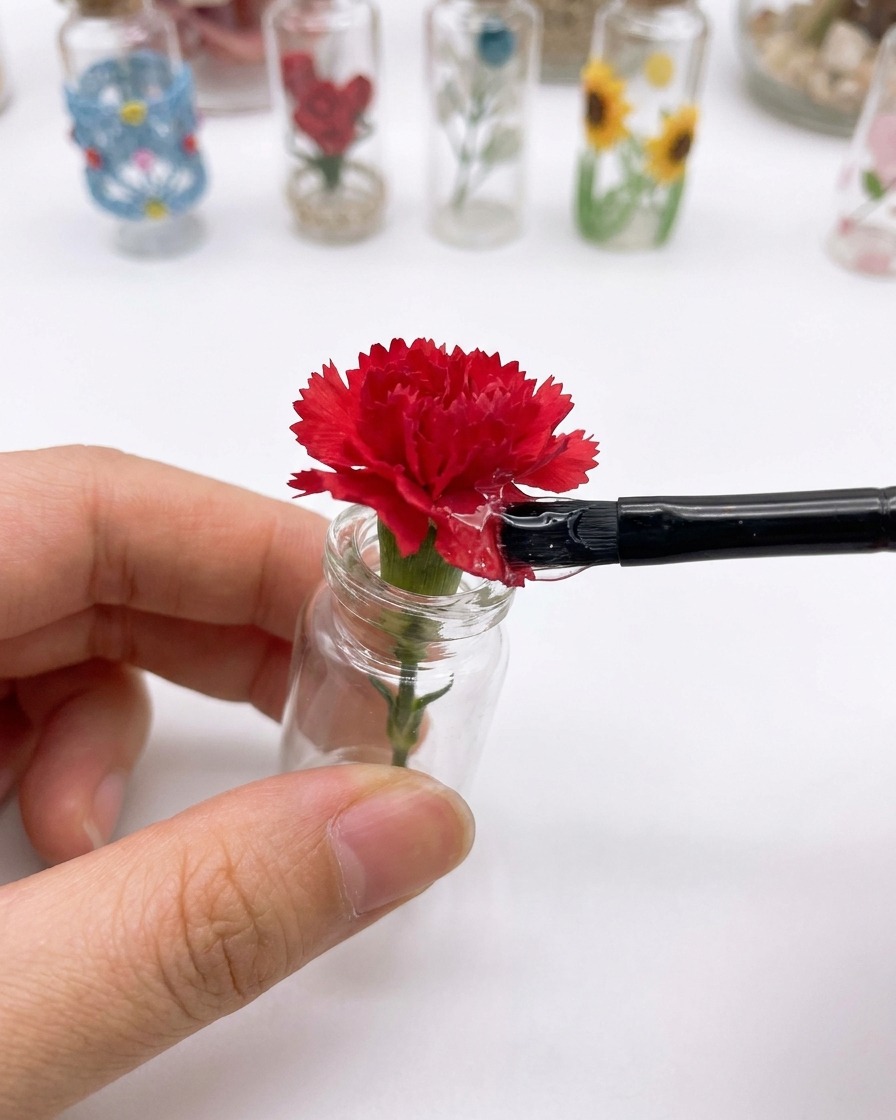 Un pincel pequeño aplicando un top coat transparente sobre la superficie de una flor de clavel roja dentro de un pequeño dije de vidrio para uñas.