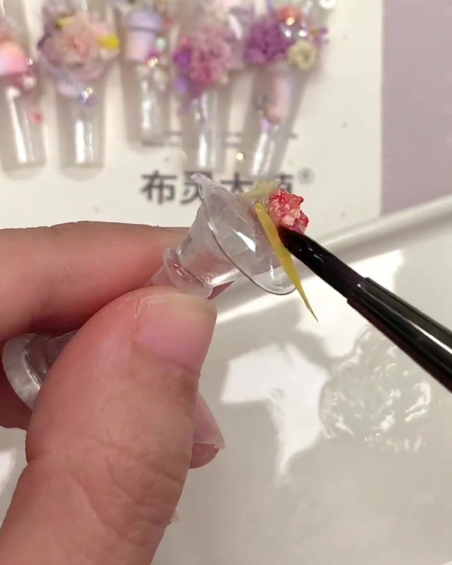 Un pincel de uñas fino aplicando top coat transparente debajo de una pequeña pieza de flor seca amarilla.