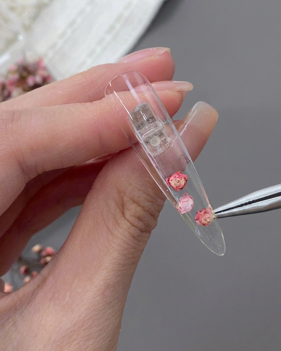 Pinzas de precisión colocando una pequeña flor seca junto a la rosa central dentro de la uña stiletto.