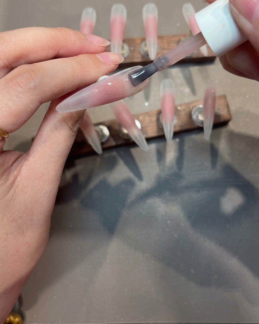 Un pincel aplicando una capa uniforme de esmalte en gel nude sobre una punta de uña con un centro rosado.