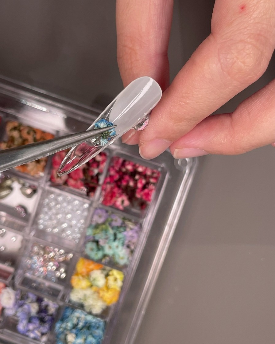 Pinzas colocando una pequeña flor azul seca en la parte inferior de un tip de uña transparente.