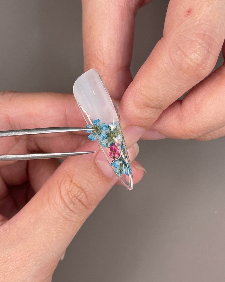 Primer plano de pedrería transparente siendo colocada entre flores secas en la parte inferior de un tip de uña.