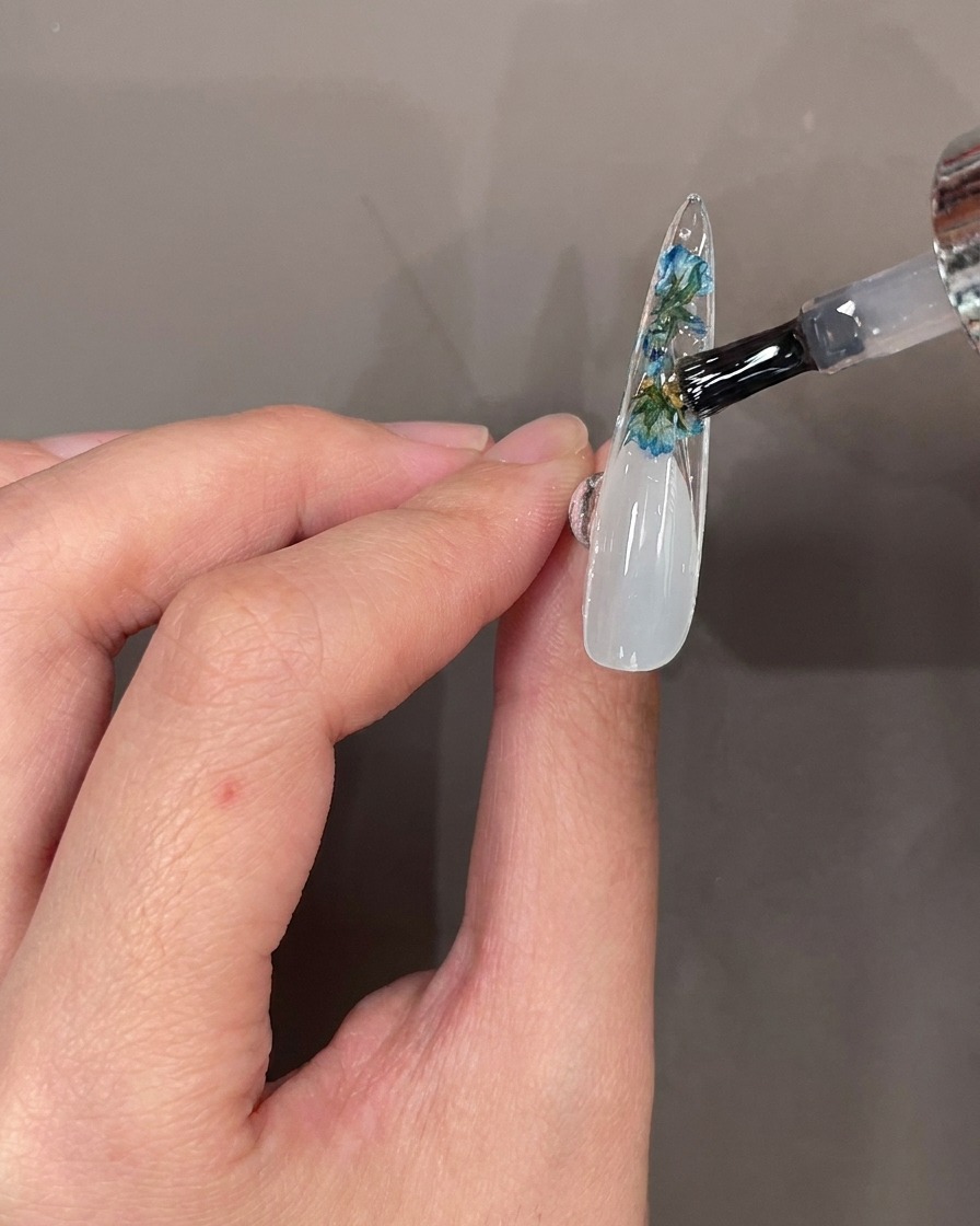 Un pincel de uñas aplicando top coat transparente sobre flores y piedras en la parte inferior de un tip de uña.