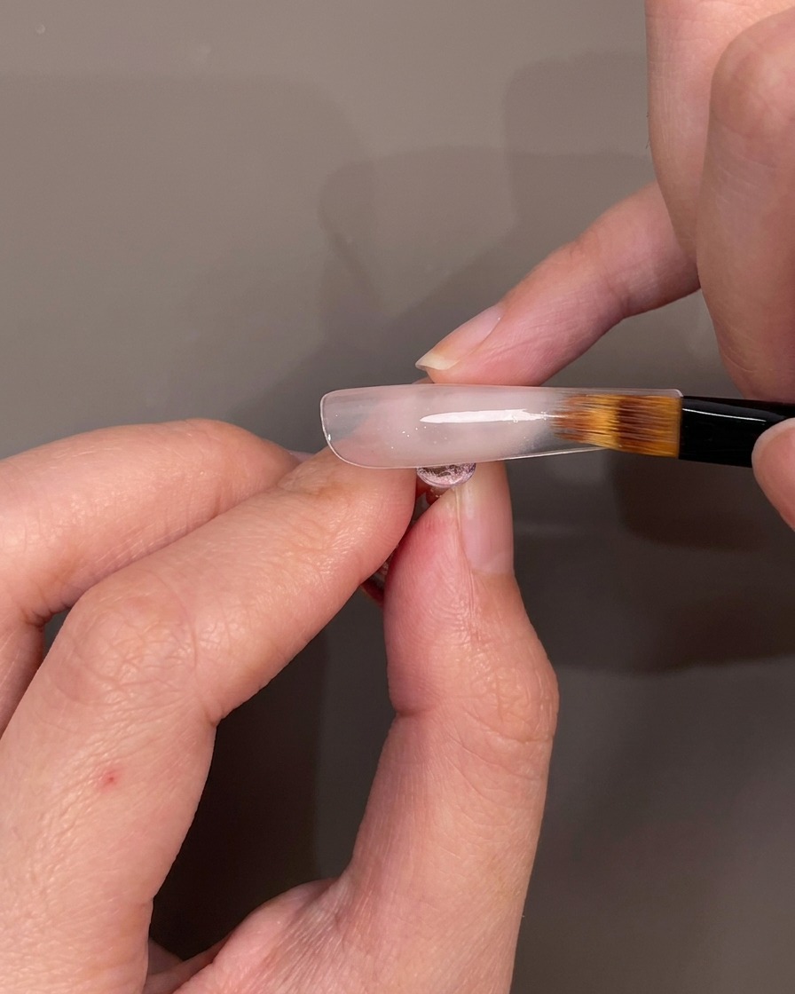 Un pincel difuminando esmalte de gel nude hacia abajo en un tip de uña para crear un efecto ombré.