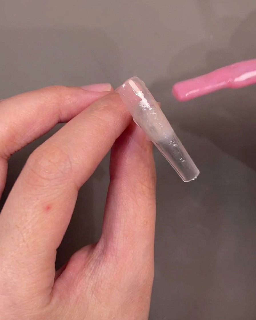 Un pincel aplicando esmalte de gel rosa transparente diagonalmente a través de un tip de uña.