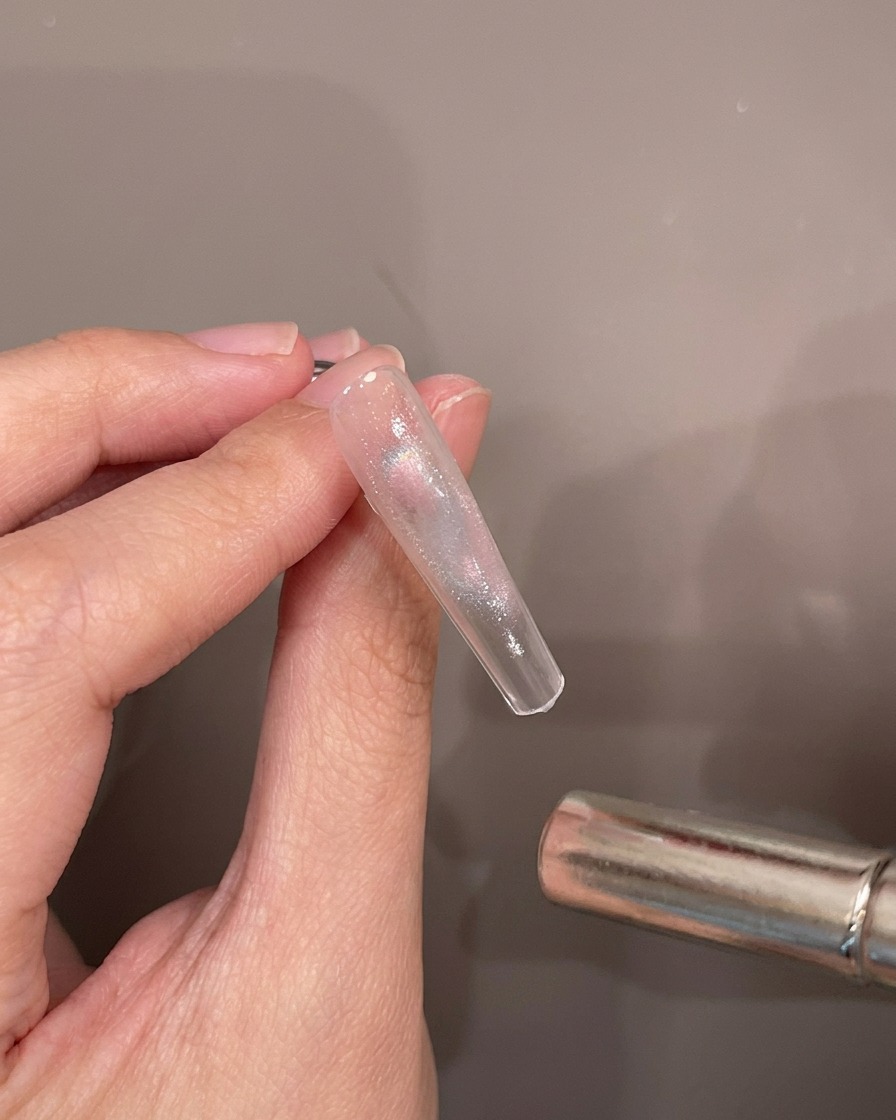 Una mano sosteniendo un tip de uña transparente mientras una varita magnética cilíndrica se cierne sobre el gel cat-eye húmedo para crear un efecto de brillo.