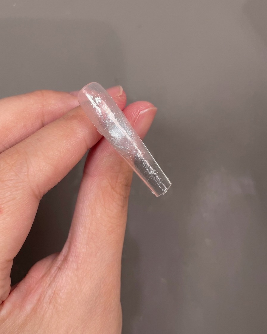 La parte inferior de un tip de uña transparente con una pequeña flor seca azul siendo colocada cuidadosamente en una capa de gel húmedo.