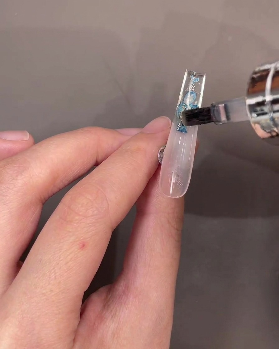 Un pincel aplicando una capa de top coat transparente sobre las flores secas en la parte inferior de una uña transparente.