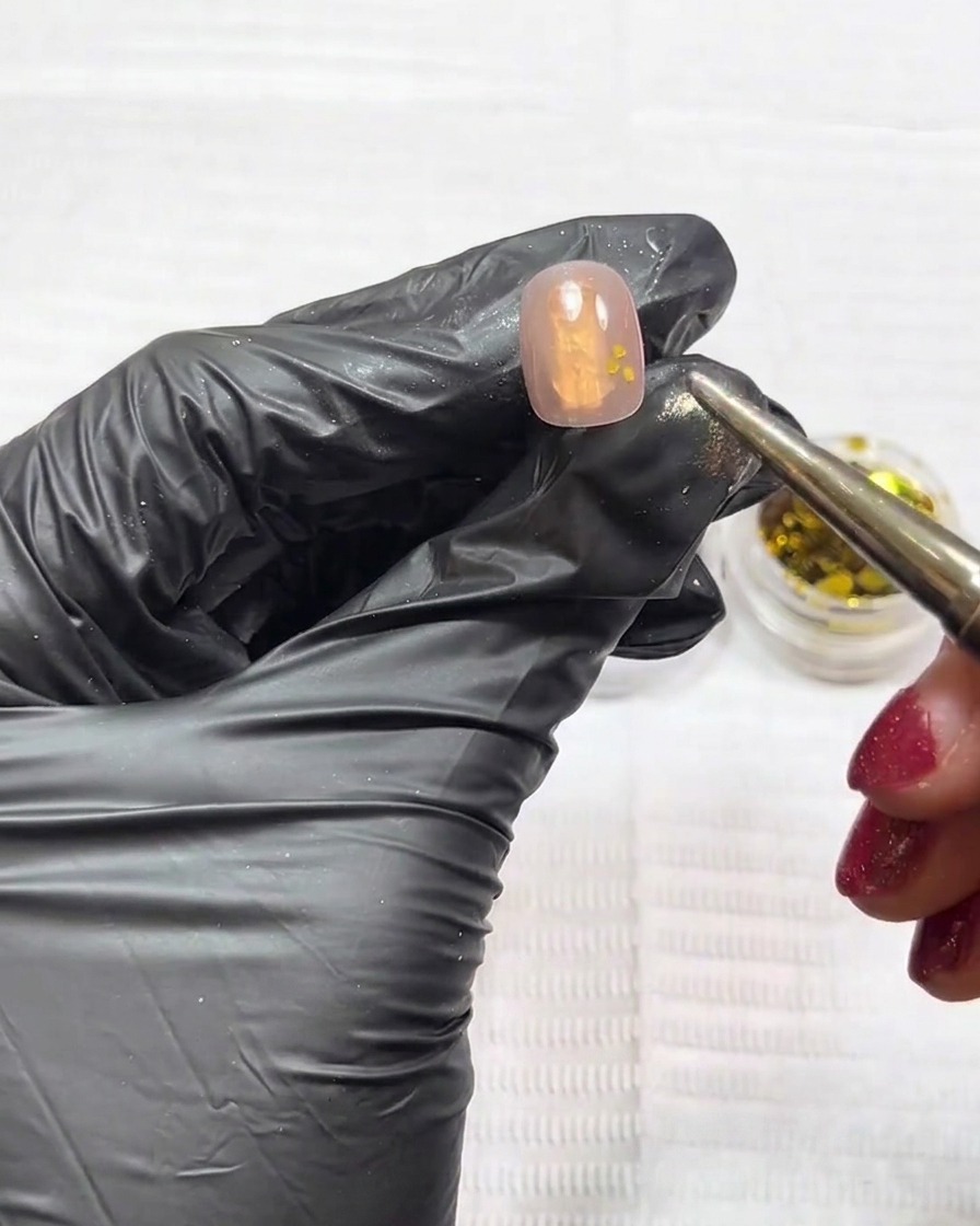 Primer plano de una uña mostrando purpurina dorada y plateada siendo colocada con una herramienta sobre una base nude.
