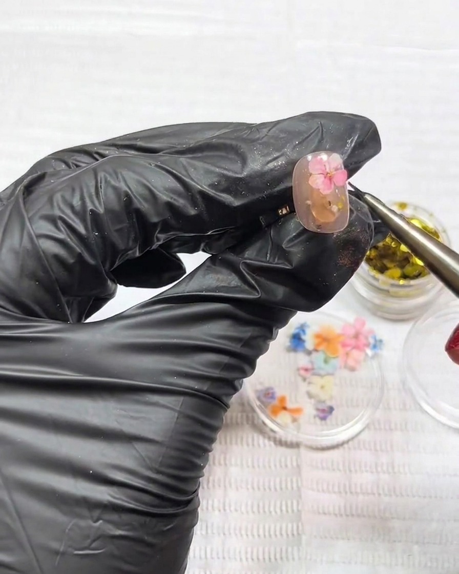 Unas pinzas colocando una pequeña flor seca rosa plana sobre la superficie de una uña.