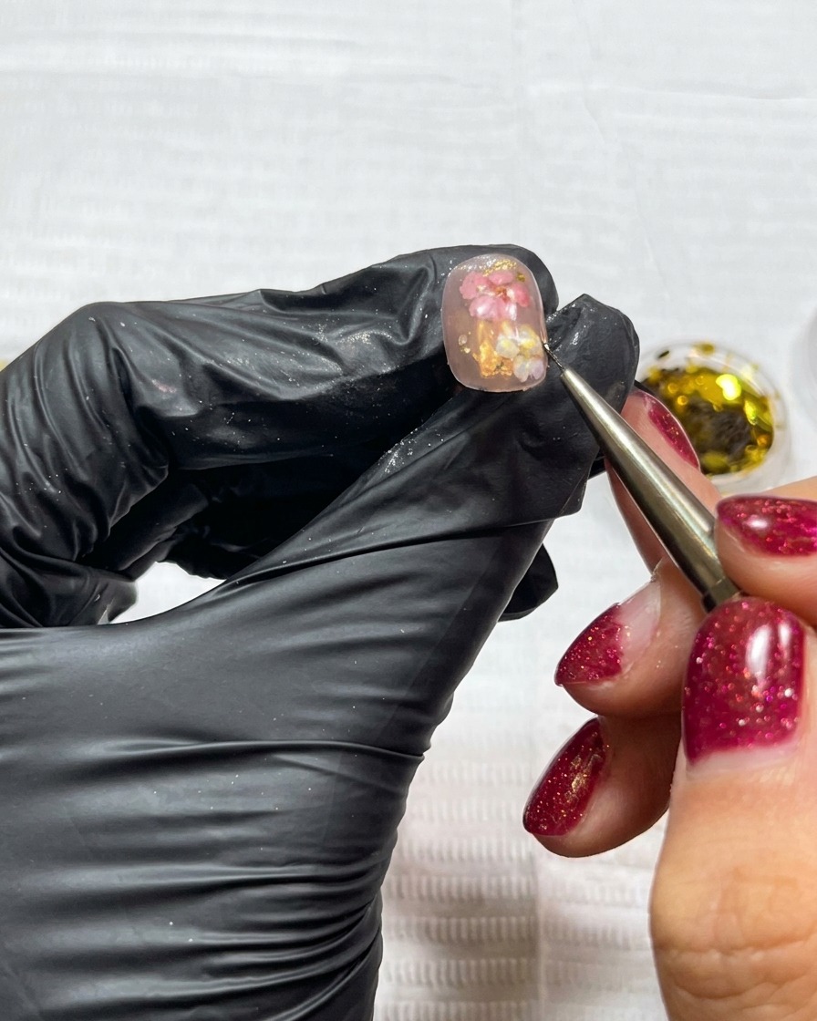 Un pincel de detalle aplicando pequeños copos irregulares de pan de oro a una uña decorada con flores secas.