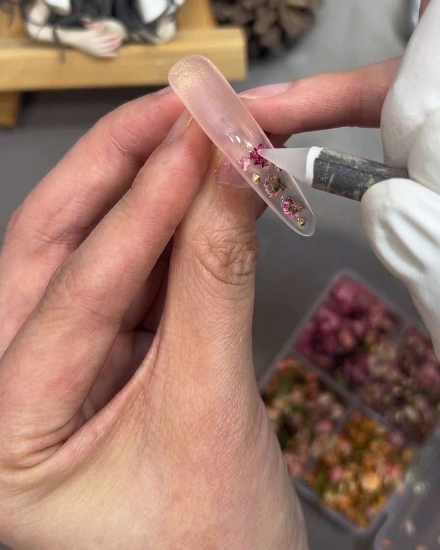 Primer plano de una mano utilizando una herramienta para colocar pequeñas perlas doradas y pedrería rosa sobre la uña.