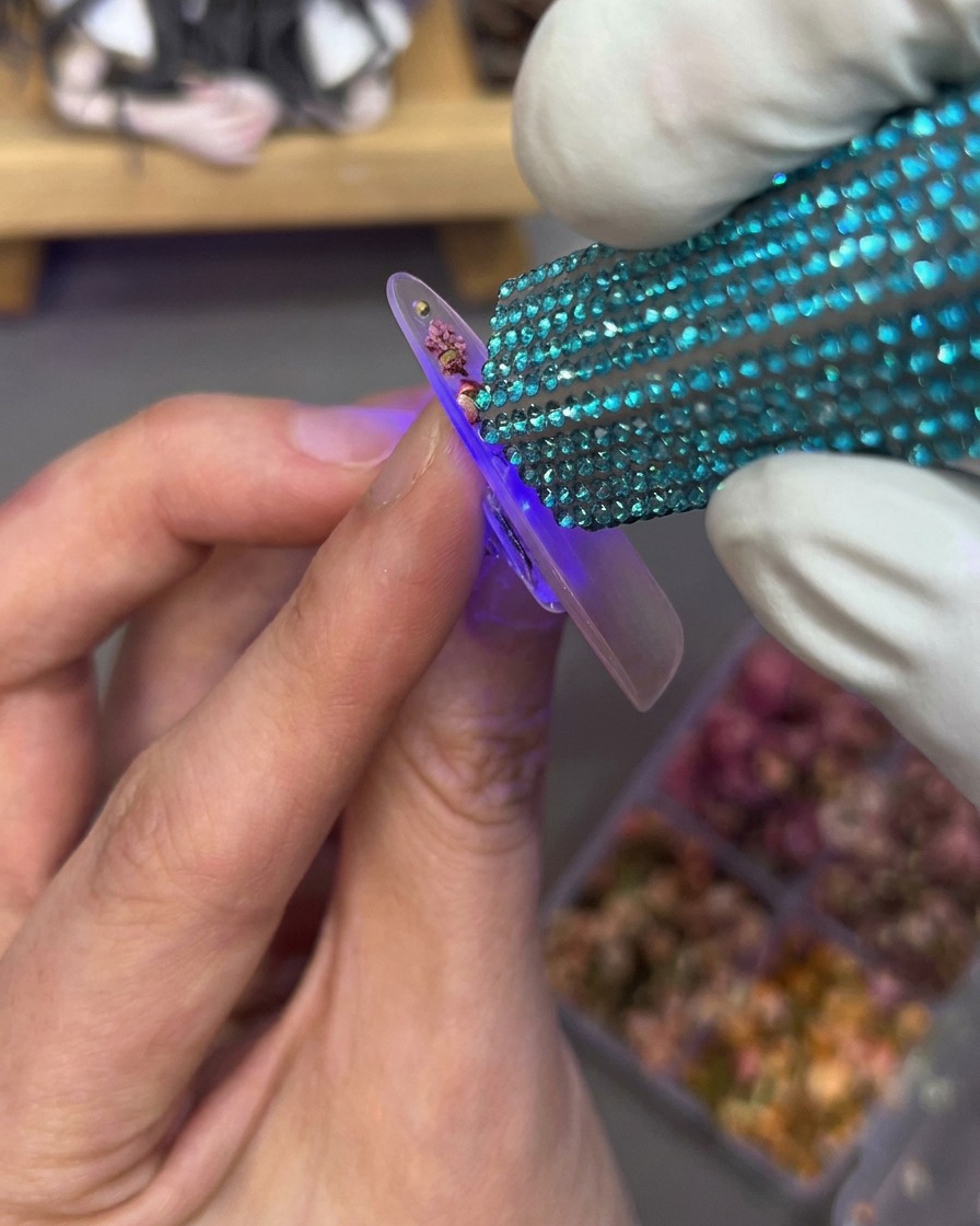 Una mano sosteniendo una mini lámpara UV sobre la uña que tiene flores secas y pedrería integradas.